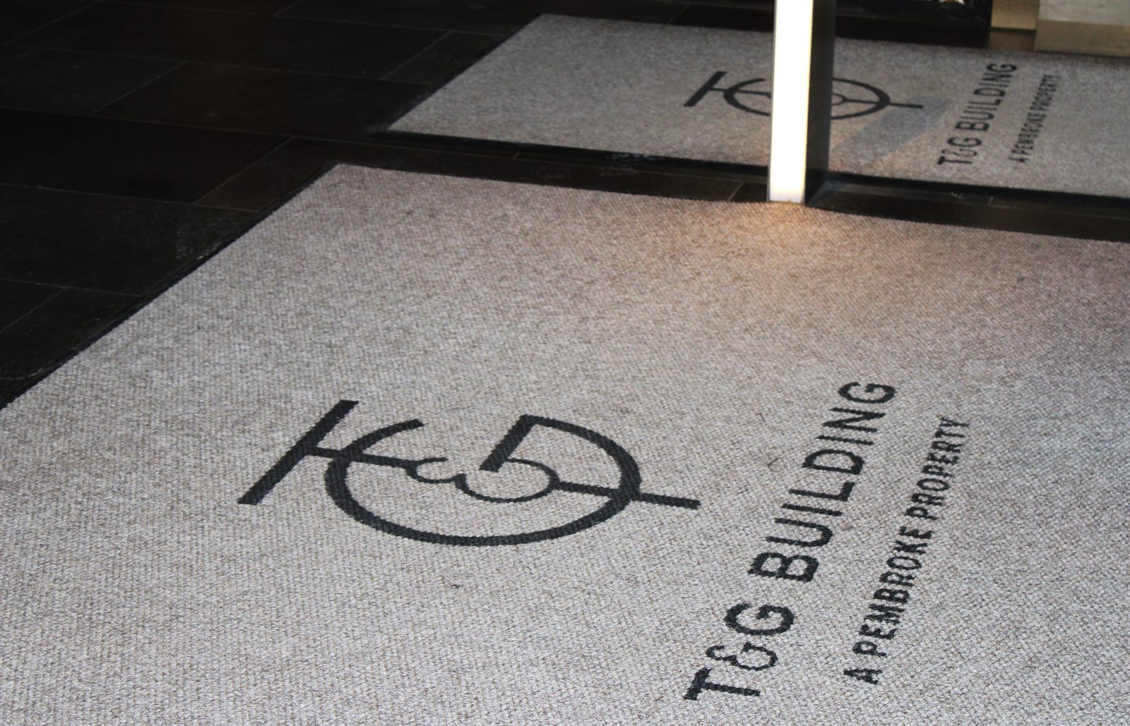 Superguard Logo Inlay mat with the T&G Building logo cut into a natural beige carpet base at a building entrance